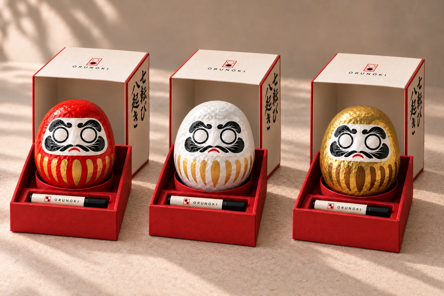 Three open gift boxes in a row, each holding one Daruma with blank eyes, in red, white and gold, with a marker pen in front of each doll.